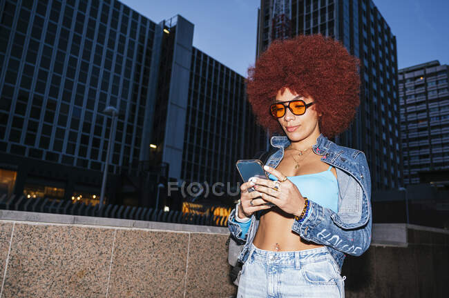 Atractiva hembra con peinado afro y mensajes de texto de ropa de moda en el teléfono celular mientras está de pie en la calle con edificios modernos en el tiempo de la noche - foto de stock
