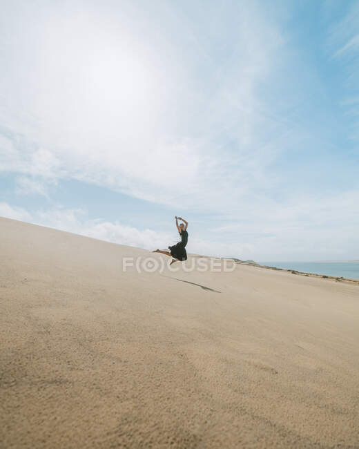 Vista lateral de cuerpo completo de bailarina bailarina descalza saltando con las manos juntas y las piernas hacia atrás en el desierto de arena - foto de stock