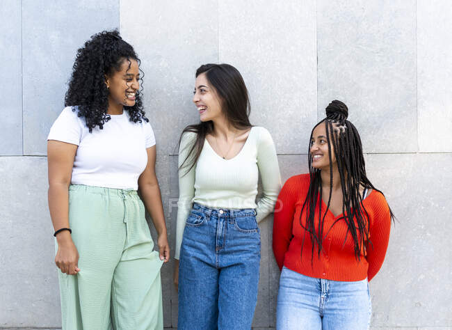 Счастливые разнообразные девушки в модной одежде стоя и смеясь во время общения рядом с серой стеной на улице днем — стоковое фото