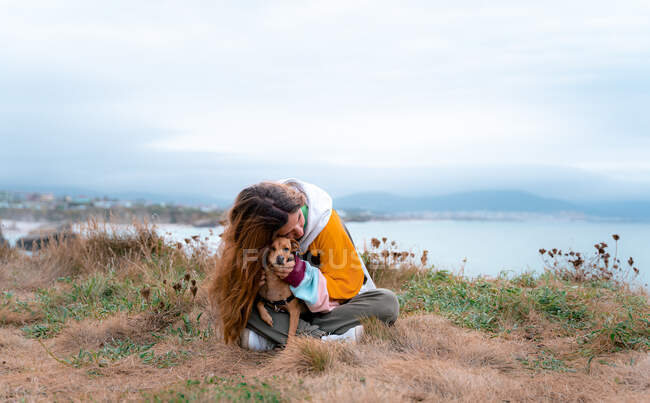 Щаслива жінка - мандрівниця обіймає гарного цуценя, сидячи зі схрещеними ногами на лузі у високогір 