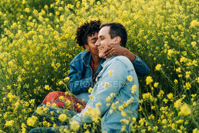 Любляча молода багаторасова пара в повсякденному одязі цілується, сидячи в пишному квітучому лузі під час романтичного побачення в сонячний день — стокове фото
