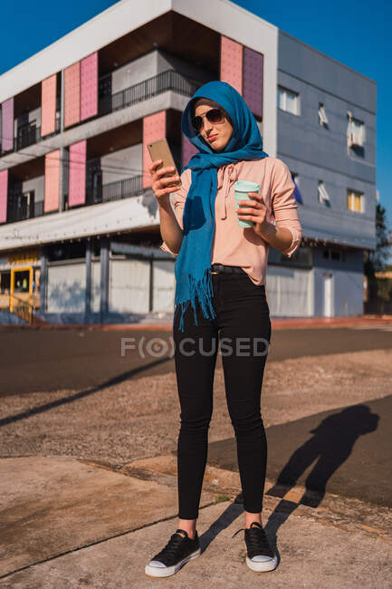 Mujer árabe de moda en el hiyab tradicional de pie con café para ir en la calle y mensajería en el teléfono celular en el día soleado en la ciudad - foto de stock