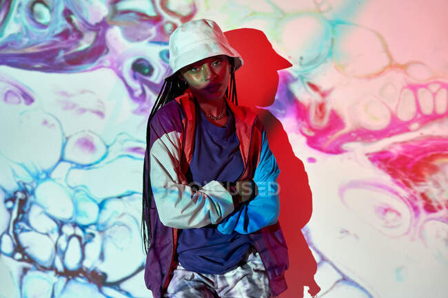 Young Dominican female teenager in trendy outfit and hat standing near white wall with creative abstract projections and looking at camera while with arms crossed — Stock Photo