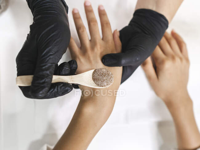 Top view of crop unrecognizable manicurist in gloves with wooden spoon with scrub above hands of client at table in salon — Stock Photo