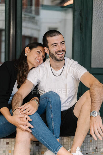 Contenuto giovane coppia etnica con i capelli scuri in abiti casual sorridente e guardando lontano mentre seduti insieme sulle scale — Foto stock