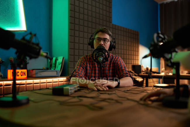 Jeune homme barbu millénariste en lunettes et écouteurs parlant en micro tout en enregistrant podcast en studio sombre — Photo de stock