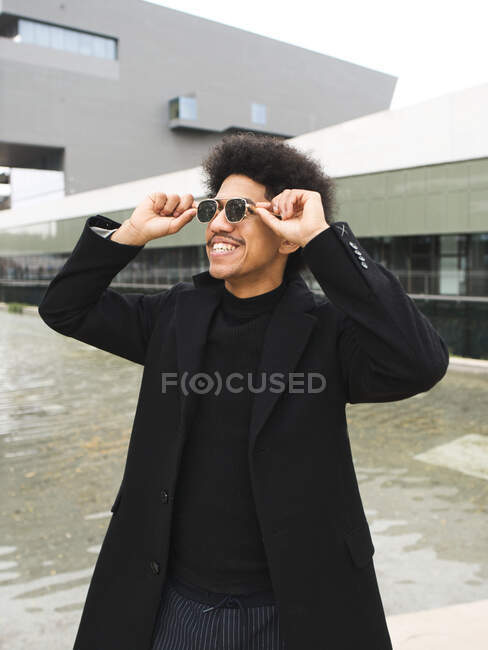 Cheerful young African American male millennial with curly dark hair adjusting sunglasses and looking away while standing near modern building on city square — Stock Photo