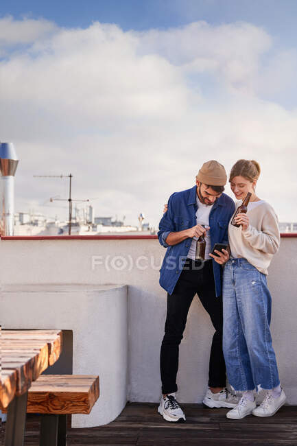 Homme barbu souriant avec bouteille de bière étreignant petite amie positive défilant téléphone mobile sur le balcon dans la journée ensoleillée — Photo de stock