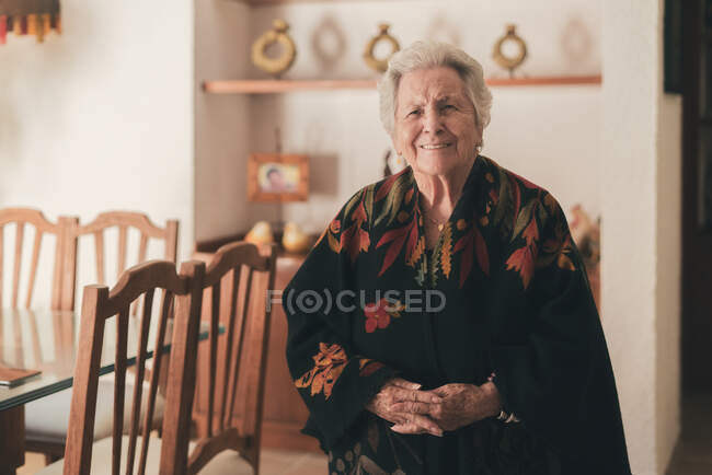 Elderly female with gray short hair and wrinkled face wearing woolen shawl in dining room and looking at camera — Stock Photo