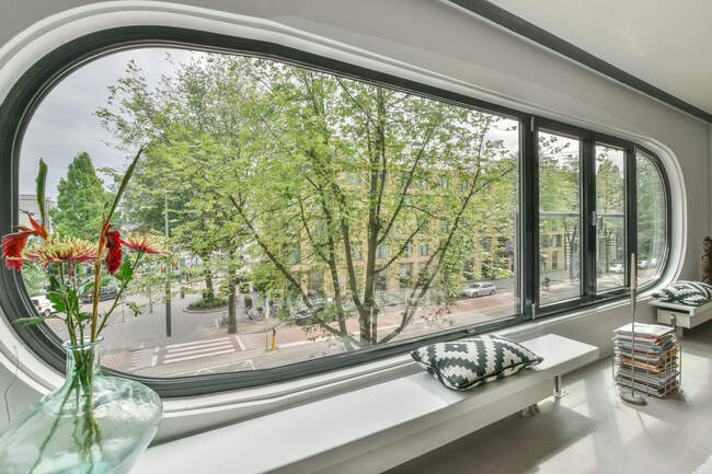 Modernes Wohnzimmerinterieur mit Sitzkissen auf Bank und blühenden Blumen in der Vase gegen ovale Fenster zu Hause — Stockfoto