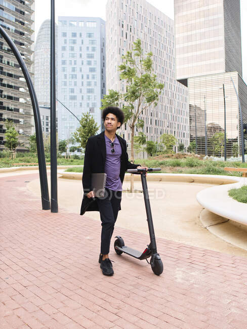 Pleine longueur de élégant jeune afro-américain millénium mâle avec ordinateur portable à la main regardant loin tout en se tenant dans le parc de la ville avec scooter électrique à proximité de différents bâtiments commerciaux modernes — Photo de stock