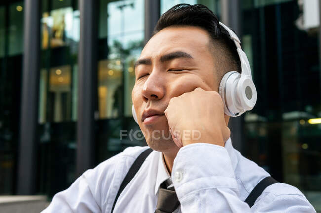 Tired young Asian male entrepreneur with closed eyes enjoying song from wireless headphones against city street — Stock Photo