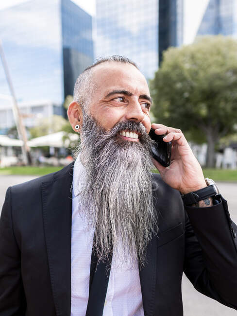 Confident smiling bearded male in classy suit having phone conversation while standing near road on street with modern buildings in city — Stock Photo