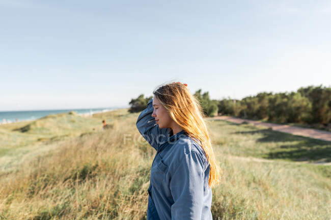 Vista lateral de la joven mujer reflexiva en traje casual de pie en el prado herboso en verano con los ojos cerrados - foto de stock