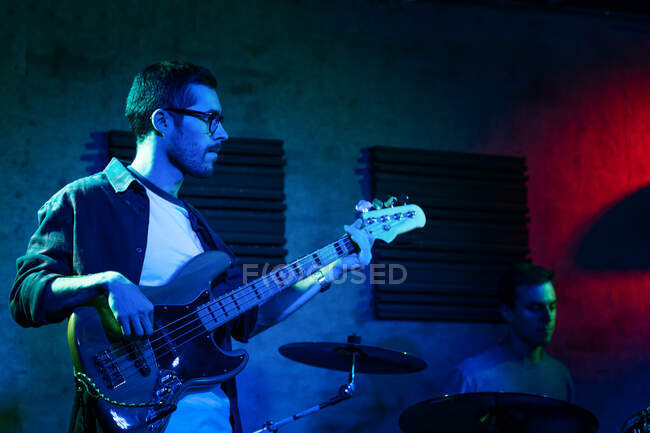 Серйозні юнаки грають на барабанах і бас-гітарі під час виступу в легкому клубі з неоновим освітленням — стокове фото