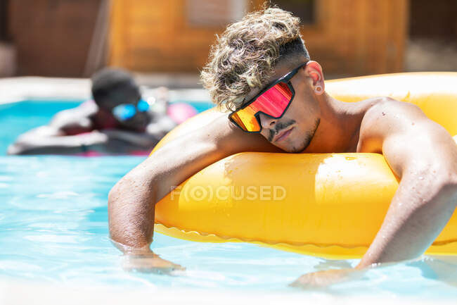 Багаторасові друзі чоловічої статі в басейнах лежать на надувному кільці і матраці під час сонячних ванн в басейні в сонячний літній день — стокове фото