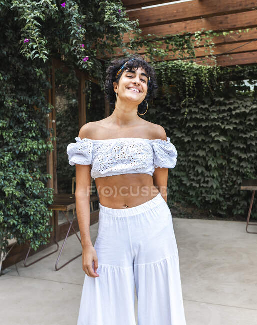 Delighted ethnic female in stylish white clothes standing in summer garden and looking at camera — Stock Photo