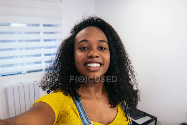 Happy African American female blogger with long curly hair in yellow t shirt and jeans overall standing with hand in pocket and taking selfie on smartphone in light room — Stock Photo