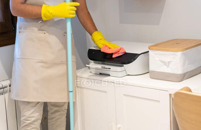 Anonyme männliche Reinigungskraft in Schürze und Gummihandschuhen waschen Boden und wischen Staub vom Drucker im Arbeitszimmer — Stockfoto