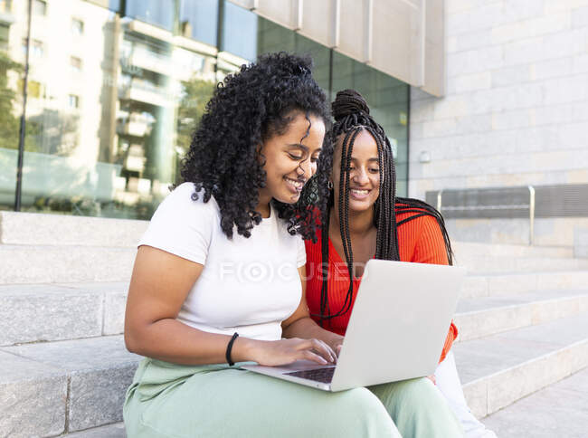 Веселі різноманітні друзі-жінки в модному одязі, сидячи на сходах і переглядаючи ноутбук на міській вулиці в літній день — стокове фото