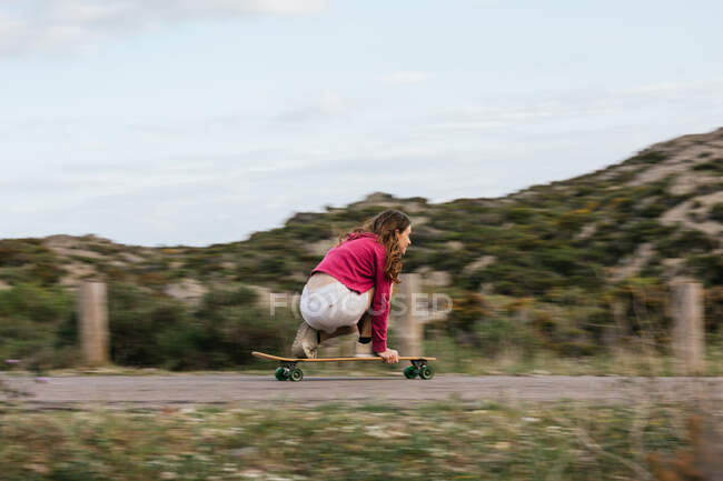 Visão lateral do corpo inteiro da jovem mulher feliz em joelheiras montando longboard na estrada de asfalto entre colinas sob céu nublado — Fotografia de Stock