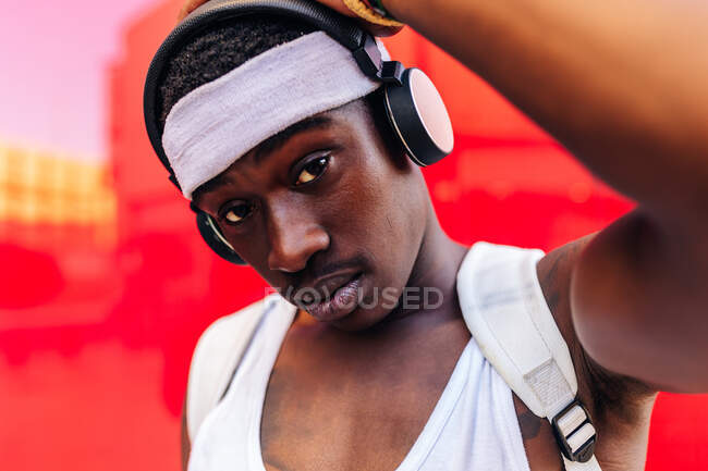 Serious African American male in wireless headphones standing against red wall in city street and looking at camera — Stock Photo