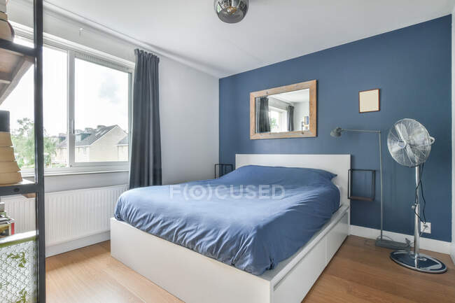 Удобная кровать с голубым одеялом размещена в стильной спальне с вентилятором и креативными декоративными элементами на стене в современной квартире — стоковое фото