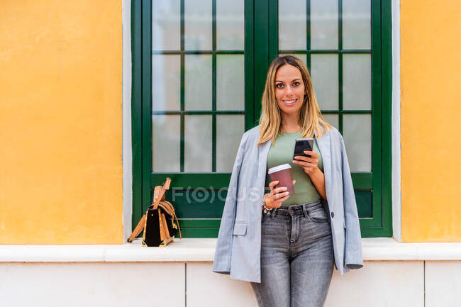 Позитивна жінка в стильному вбранні з кавою, що виймається, дивлячись на камеру, а текстові повідомлення на мобільний телефон біля будівлі з гаманцем — стокове фото