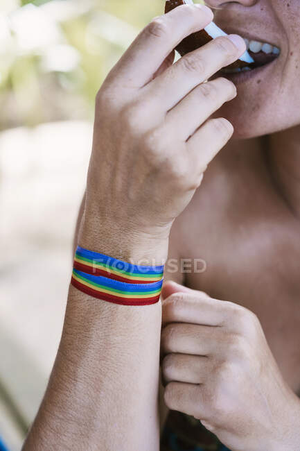 Crop unrecognizable person with rainbow ribbon on hand biting slice of fresh natural coconut against green plants in summer time — Stock Photo
