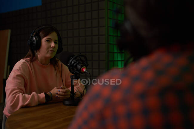 Female radio host in casual clothes and headphones while recording podcast in modern studio with crop unrecognizable colleague — Stock Photo