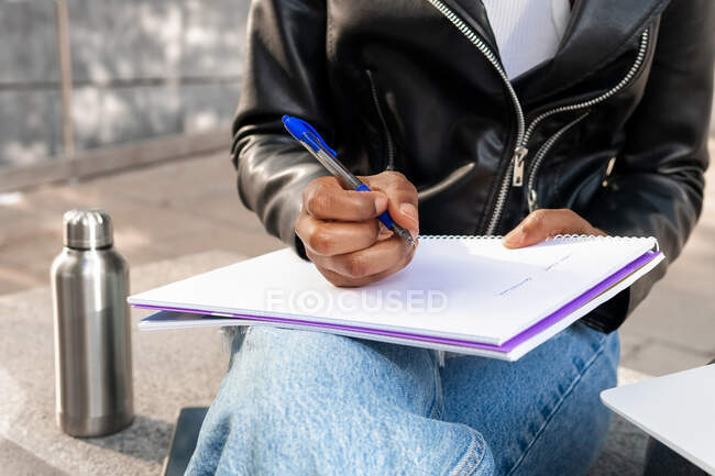 Anonymous African American female in leather jacket taking notes in notebook on street near building in city — Stock Photo