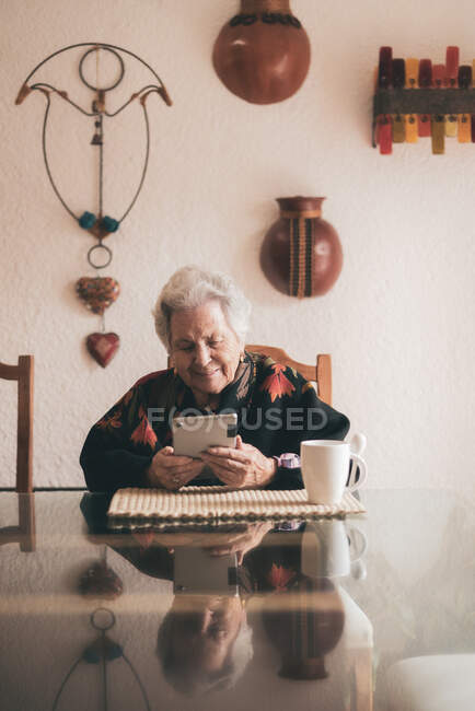Donna anziana sorridente che indossa vestiti caldi seduta a tavola con tablet e tazza di tè guardando lo schermo — Foto stock
