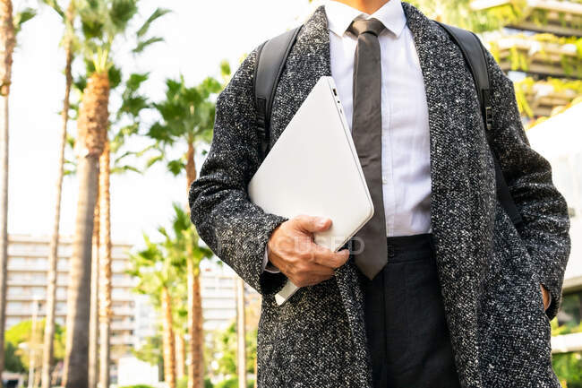 Cropped unrecognizable male entrepreneur with rucksack and netbook looking away against modern urban buildings and palm trees — Stock Photo