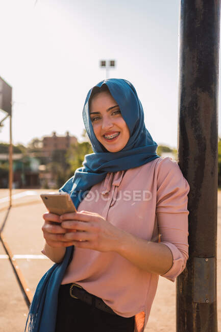 Позитивная молодая арабская женщина в хиджабе, просматривающая мобильный телефон, стоя возле столба на городской улице и глядя в камеру — стоковое фото