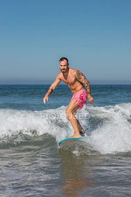 Uomo attivo in pantaloncini da bagno in piedi sulla tavola da surf mentre fa surf nel mare ondulato nella località tropicale nella soleggiata giornata estiva — Foto stock