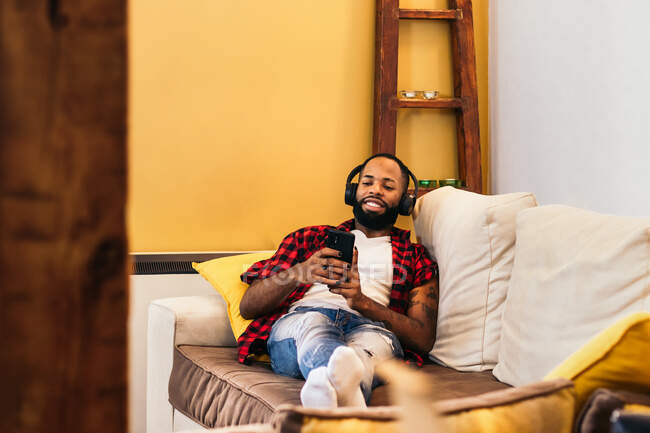 Smiling tattooed ethnic male with manicure chatting on cellphone while lying with crossed legs on couch at home — Stock Photo
