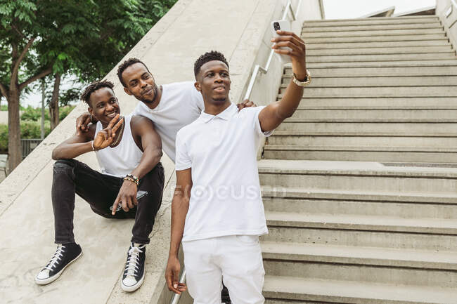 Positive African American man embracing friend showing victory gesture while fellow taking selfie on cellphone — Stock Photo