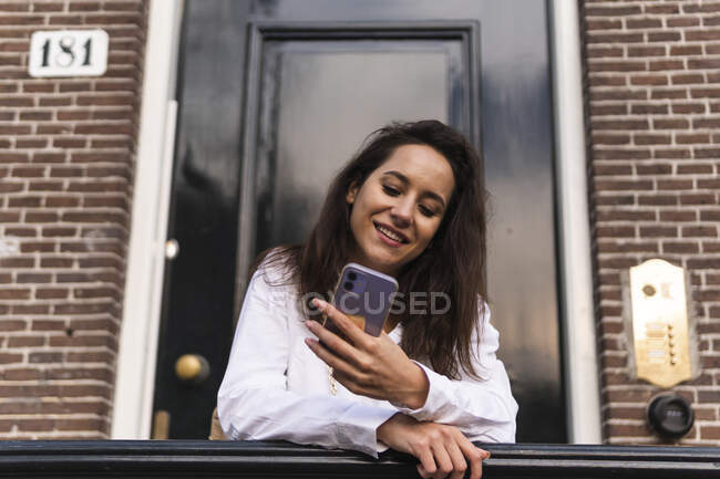 Happy young female in casual clothes standing near entrance of building and leaning on handrail while using smartphone — Stock Photo