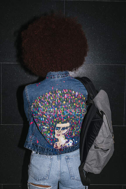 Back view of anonymous female with Afro hairstyle and stylish outfit standing near black wall with backpack on street in evening time — Stock Photo