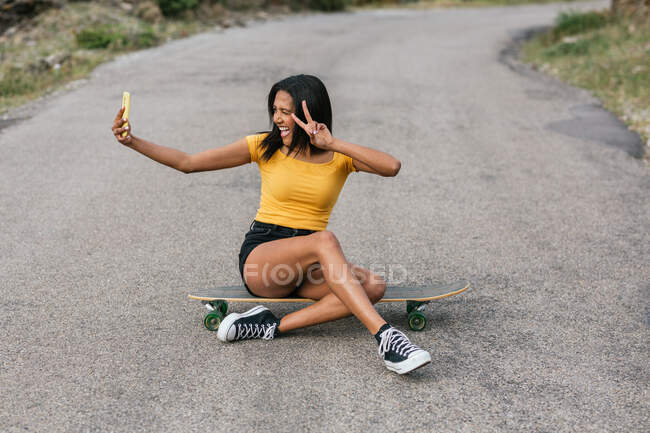 Corps complet d'une heureuse femme ethnique assise sur un longboard avec des jambes croisées tout en montrant un geste de paix et en prenant un autoportrait sur un téléphone portable — Photo de stock