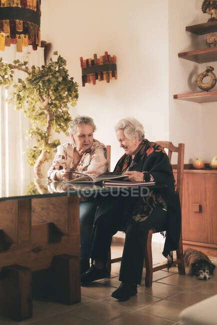 Літні сестри оглядають фотографії в фотоальбомі та обговорюють спогади, сидячи за столом вдома разом — стокове фото