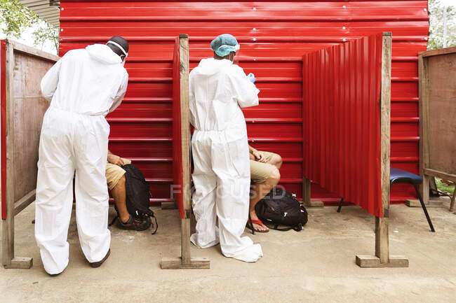 Vue arrière de médecins africains méconnaissables en uniforme de protection prélevant un échantillon de test PCR chez des voyageurs anonymes dans des cabines rouges à la campagne — Photo de stock