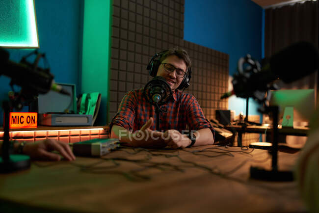 Positivo giovane barbuto maschio millenario in occhiali e cuffie sorridente e parlando in microfono durante la registrazione di podcast in studio scuro — Foto stock