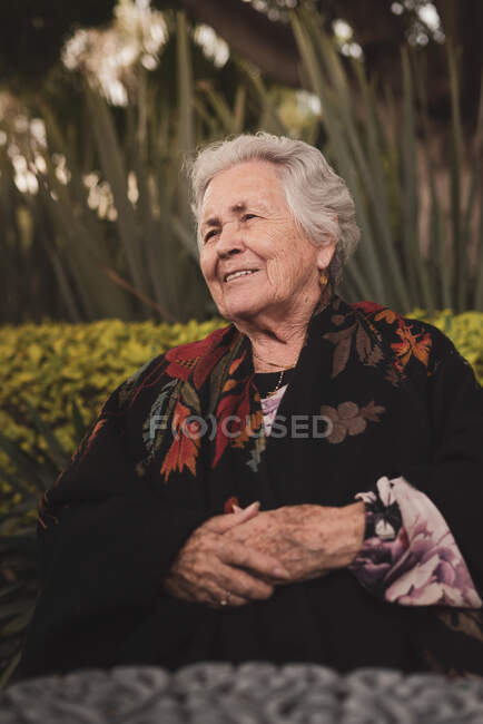 Side view of thoughtful elderly female with gray hair wrapping in plaid sitting at garden table and looking away on sunny day — Stock Photo