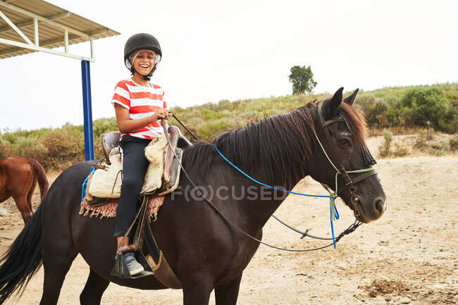 Cuerpo completo de niño sonriente en casco y ropa casual sentado a caballo en silla de montar en suelo arenoso en el rancho durante el día cerca del campo herboso - foto de stock