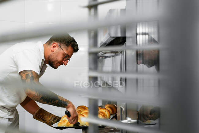 Panadero masculino con tatuajes en el brazo horneando croissants en horno metálico grande durante el trabajo en panadería - foto de stock