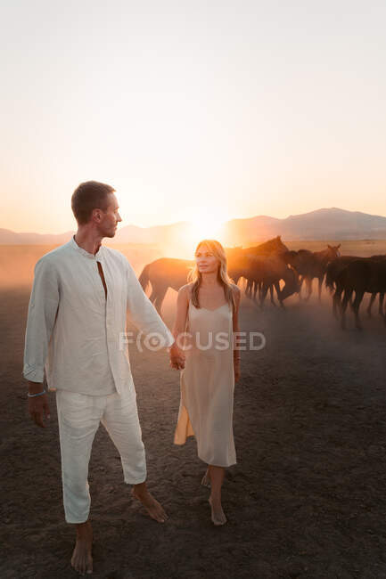 Full body of loving woman and man holding hands and looking at each other while walking in countryside in sunset — Stock Photo