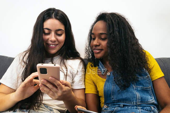 Junge Freundinnen in lässiger Kleidung lächeln, während sie zu Hause auf dem Sofa sitzen und auf dem Smartphone surfen — Stockfoto