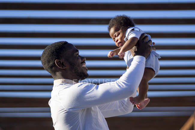 Seitenansicht eines glücklichen afroamerikanischen Vaters, der an einem sonnigen Tag ein kleines Baby auf Händen gegen eine blaue Wand auf der Straße hebt — Stockfoto