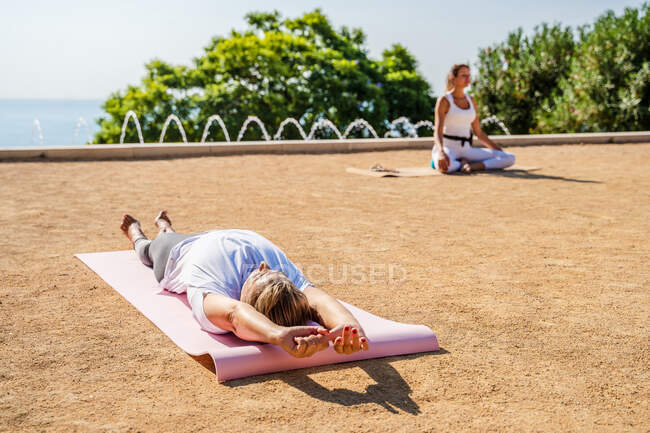 Инструктор по йоге в активной одежде сидит в позе лотоса, пока люди лежат на ковриках на земле во время Шавасаны в парке в солнечный день — стоковое фото